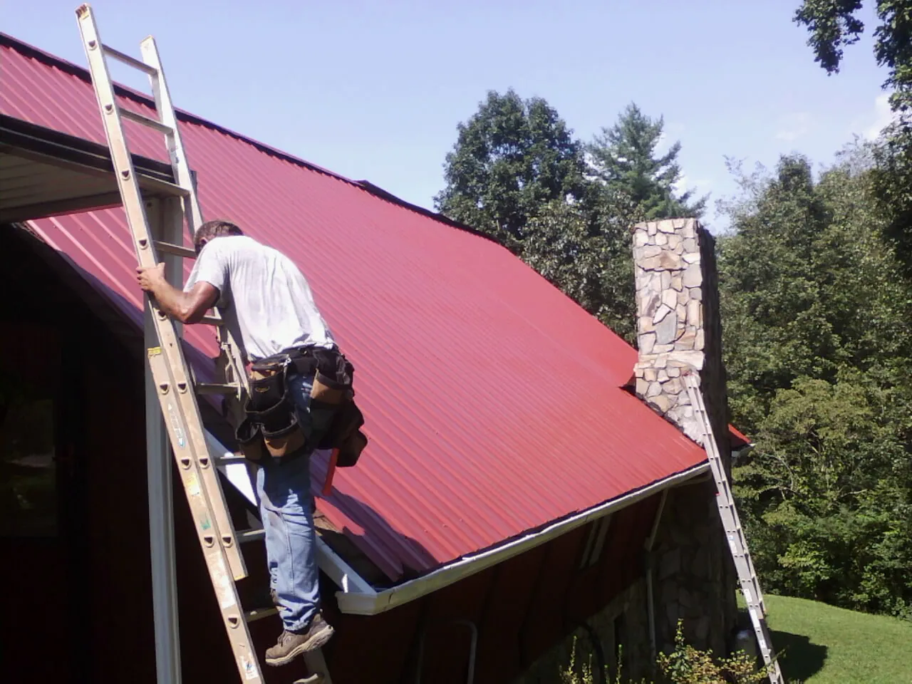 Roofer on a ladder beside a red metal roof and stone chimney — Soffit & Fascia Repair by Roofing Brothers in Eltopia